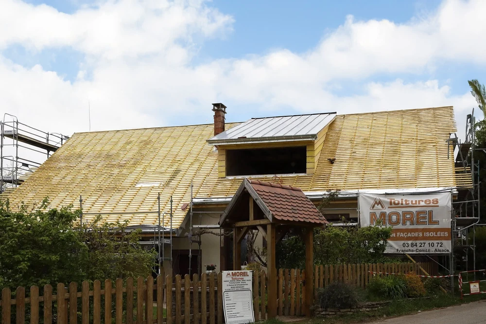 Maison contemporaine en construction avec façade mixte bois et bardage gris, toiture en tuiles métalliques avec système de fixation apparent